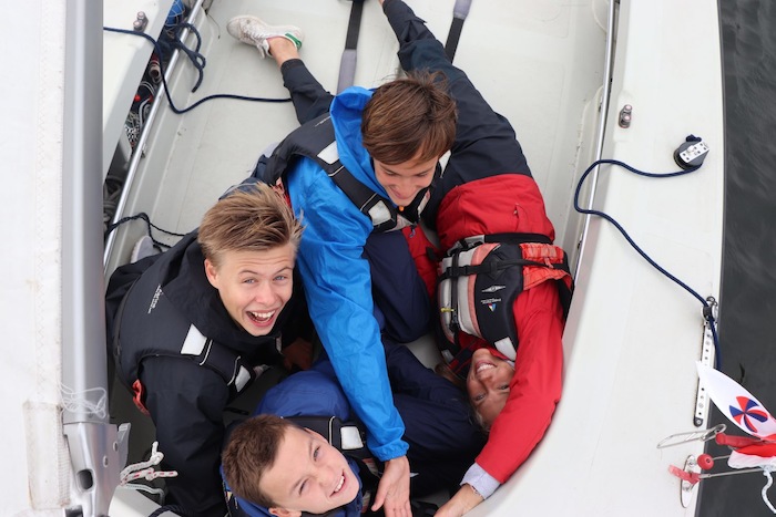 Jongeren zwaaien vanuit een zeilboot naar de camera tijdens het zeilkamp