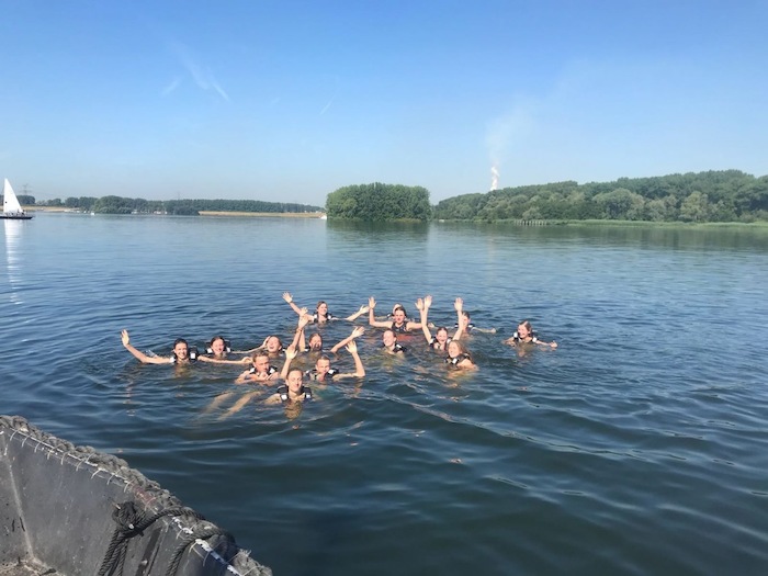 Groep jongeren zwemmen en zwaaien naar de camera op zeilkamp.
