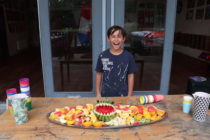 jongen lacht breeduit achter een tafel met fruitschaal tijdens zeilkamp kinderen