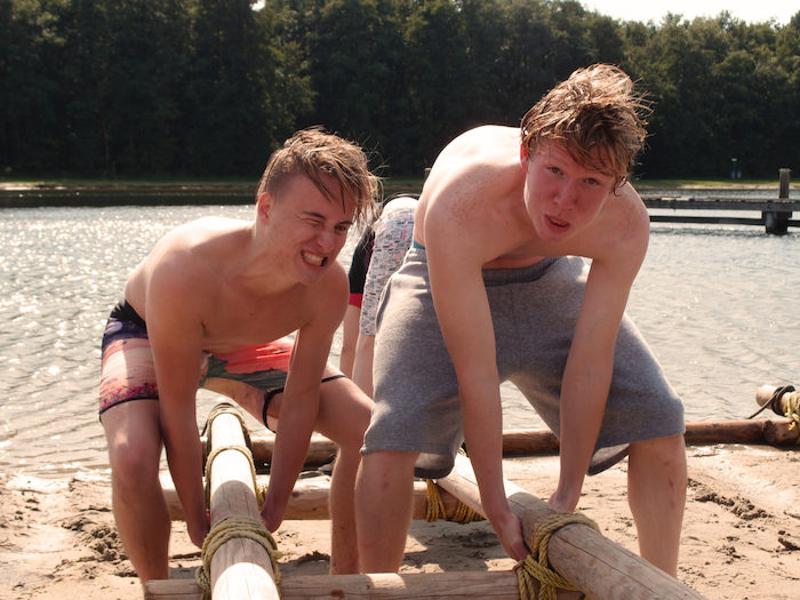 Jongens tillen balk op, kijken naar de camera op zomerkamp.