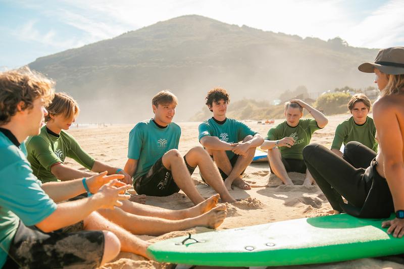 Jongeren zitten in een kringetje op het strand tijdens de surfles op het surfkamp 18 plus.