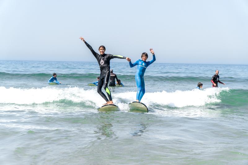 Twee deelnemers aan summer camp Portugal staan op een surfboard in de golven en houden elkaars hand vast