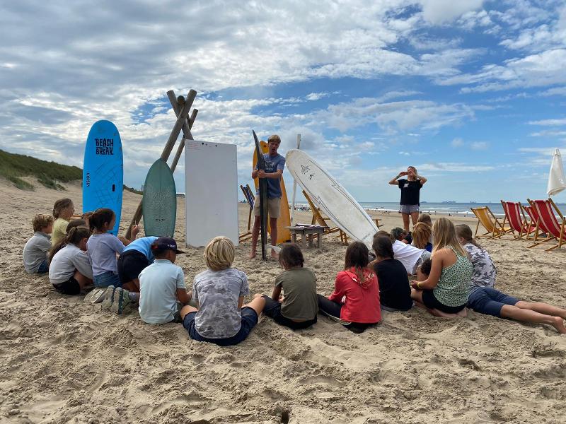 Jongeren zitten in het zand en krijgen surfles tijdens een surfkamp
