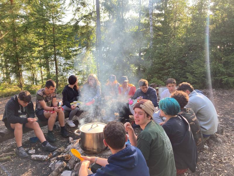 Jongeren zitten in een kring rond een pan op een kampvuur tijdens een avonturenkamp