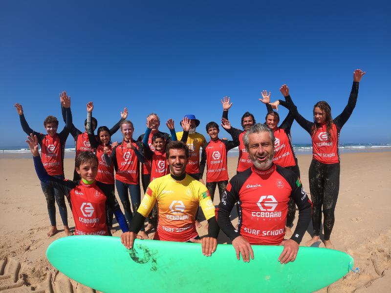 Een groep deelnemers en begeleiders van een surfkamp Portugal staan op het strand met een surfboard en juichen